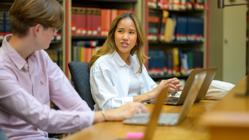 Two students in conversation with laptops in a library